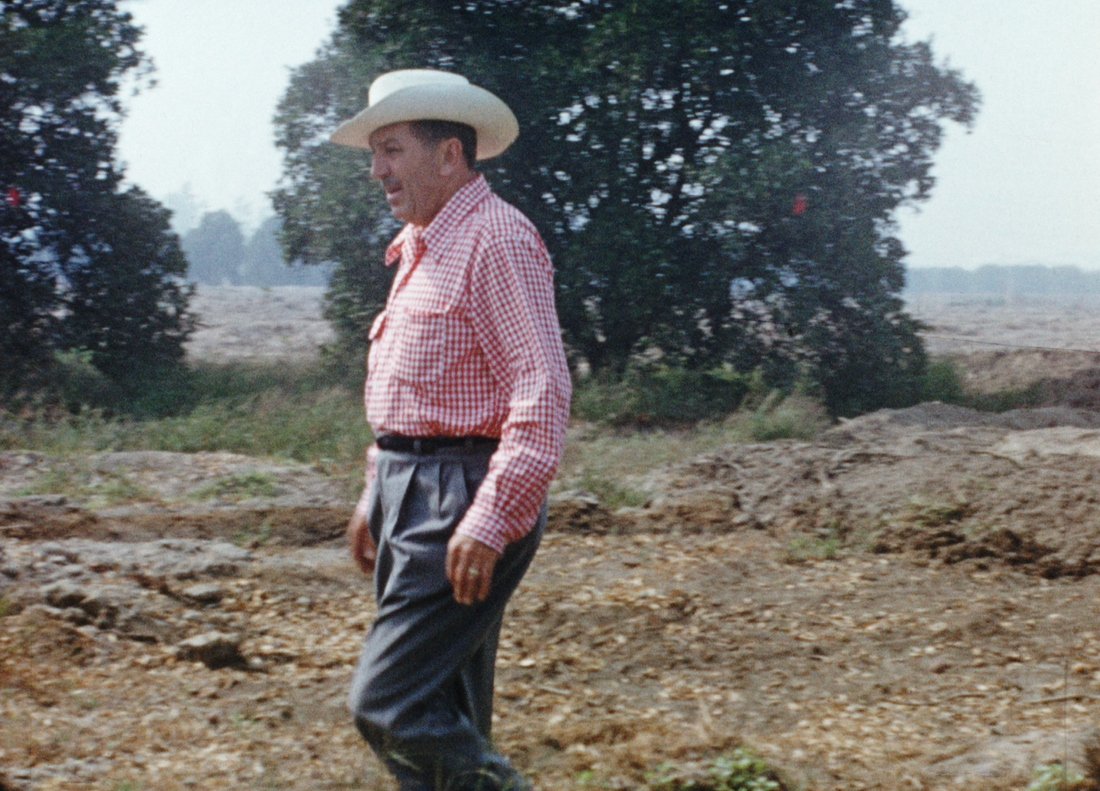Man in a white cowboy hat and red checkered shirt walks outdoors on uneven ground with trees and fields in the background.