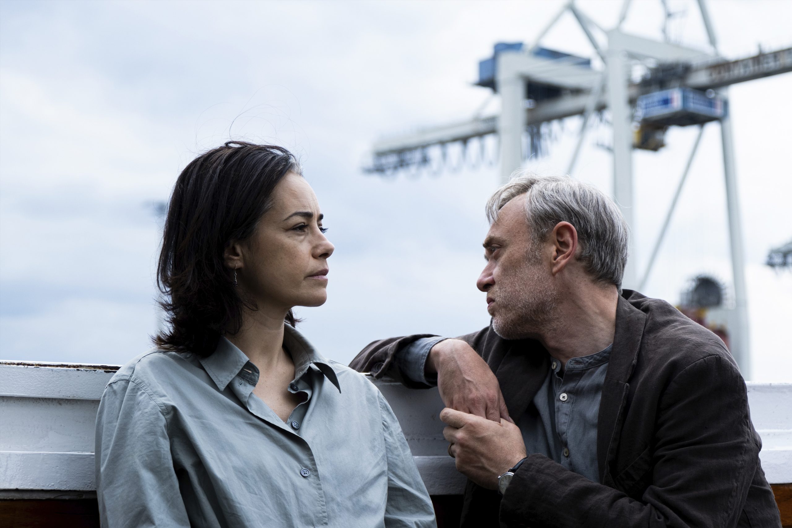 A woman and a man sit outside in front of industrial cranes, engaged in a serious conversation.