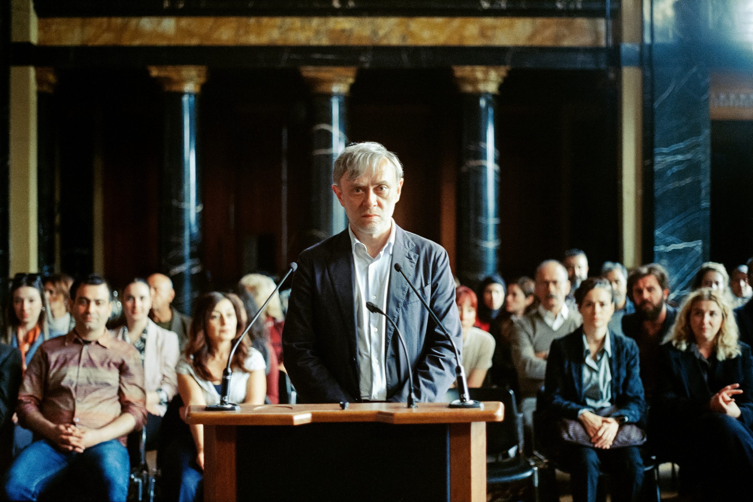 A man stands at a podium in a formal room, facing forward, with an audience seated behind him.