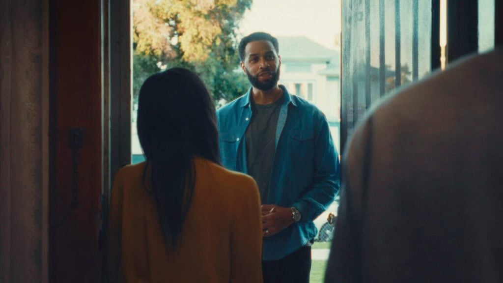 A man stands at the front door, speaking to a woman in a yellow sweater, with another person partially visible in the foreground.