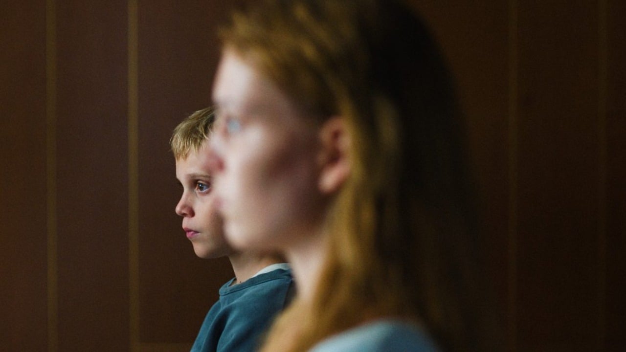 A young boy in focus sits beside an out-of-focus girl with long hair, both facing to the left against a dark, wood-paneled background.
