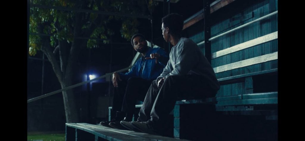 Two people sit and talk on empty bleachers at night under artificial lighting, with trees and a fence visible in the background.
