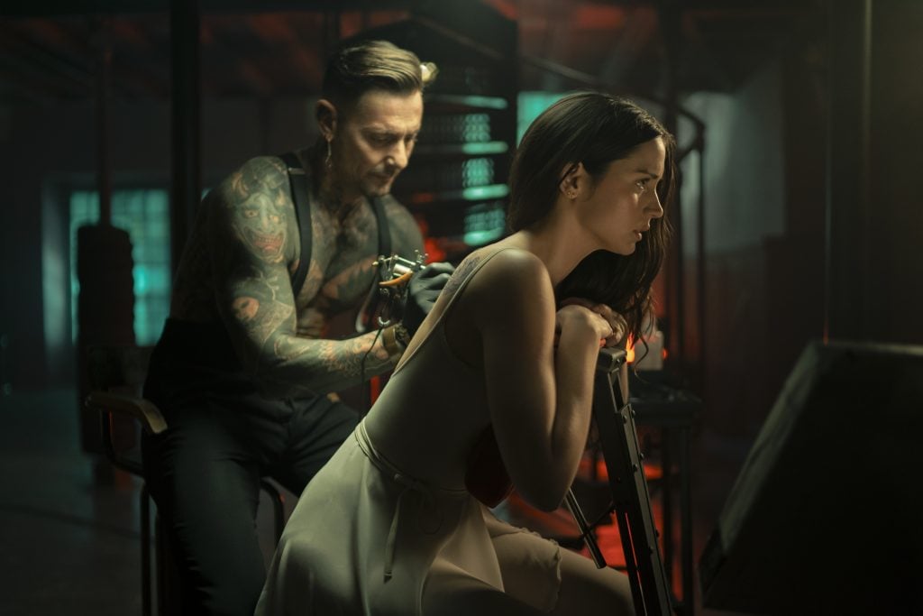 A tattoo artist with tattoos on his arms is tattooing a woman’s upper back while she sits, leaning forward in a dimly lit studio.