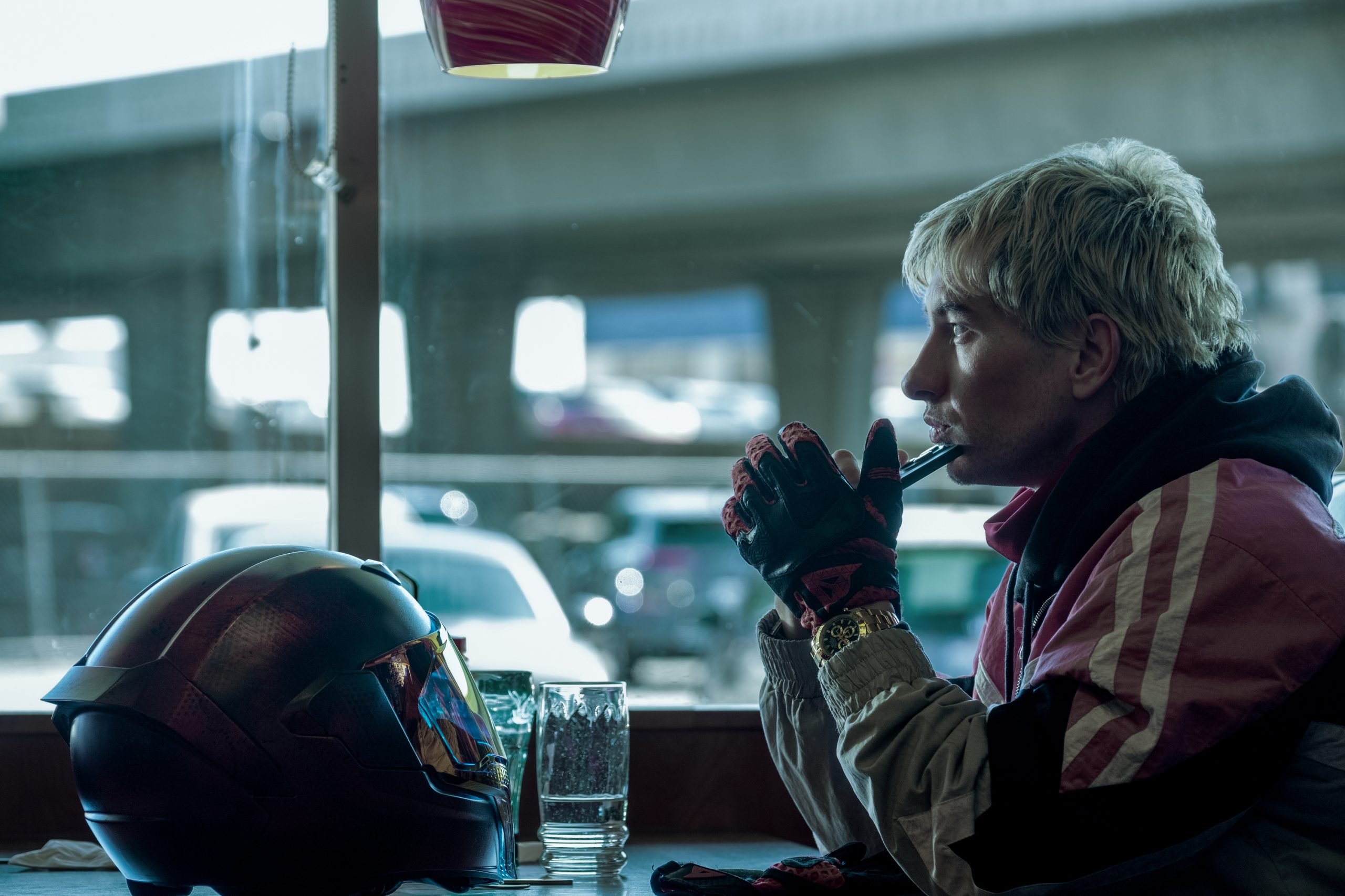 A person with blond hair and a pink jacket sits at a diner table, holding a straw near their mouth, with a motorcycle helmet and two glasses on the table.