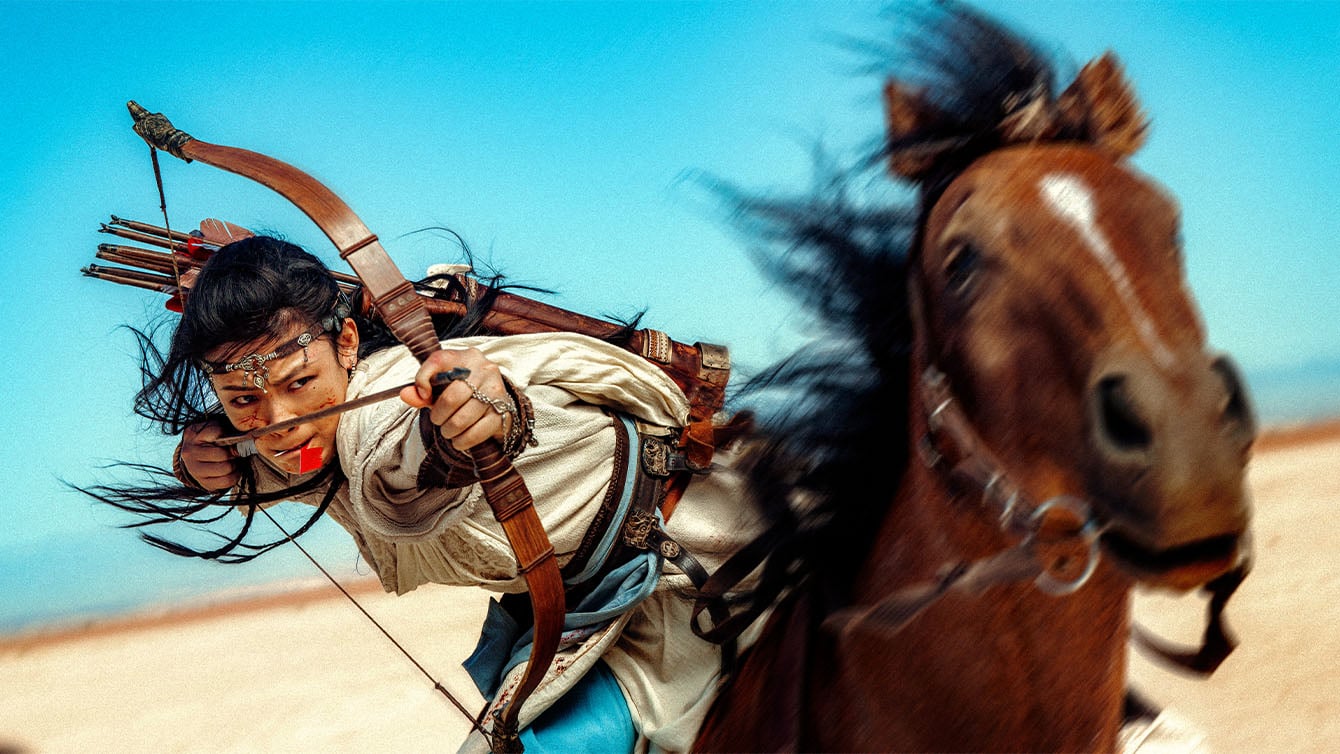 A person in traditional attire rides a galloping horse while drawing a bow and aiming an arrow, set against a clear blue sky in a desert landscape.