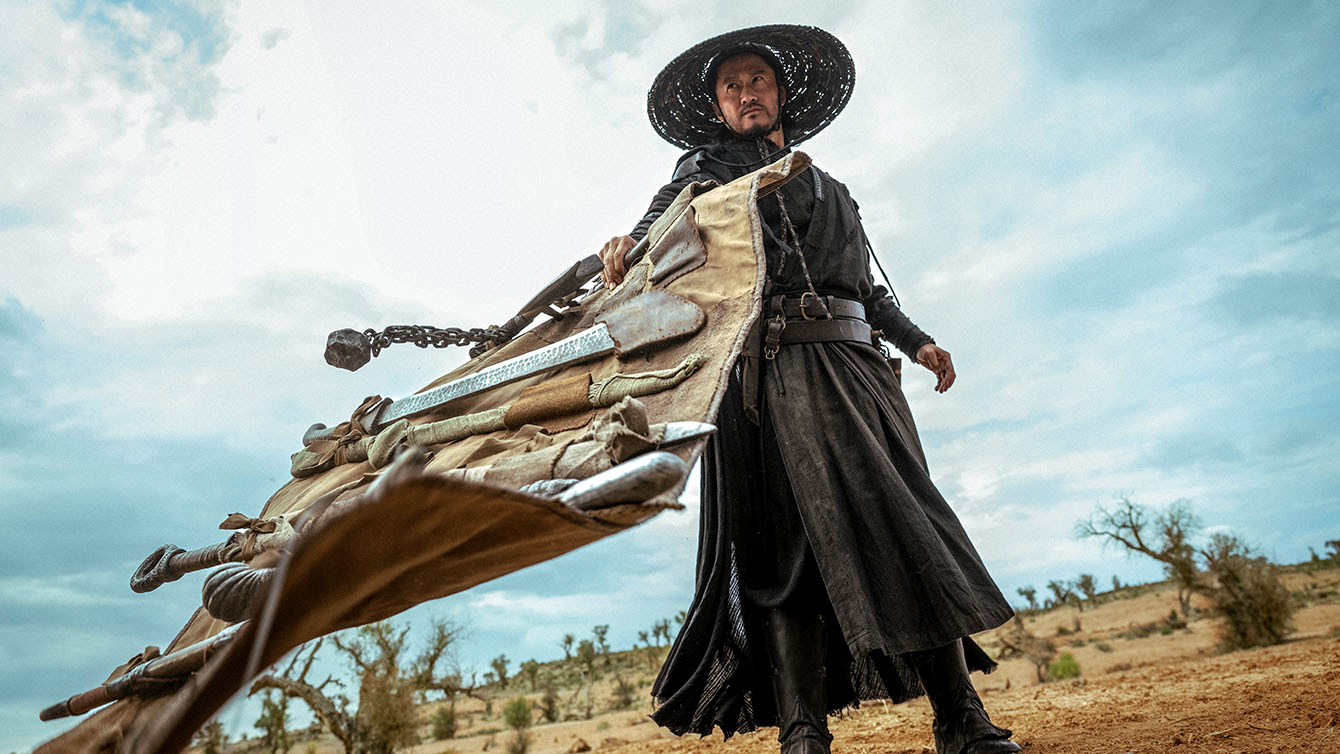 A person in dark clothing and a wide-brimmed hat stands outdoors, holding a large bundle of weapons, with a dry landscape and cloudy sky in the background.
