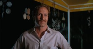 A man with blond hair and a mustache stands under a yellow canopy at night, wearing a pale button-up shirt, with hanging decorations in the background.