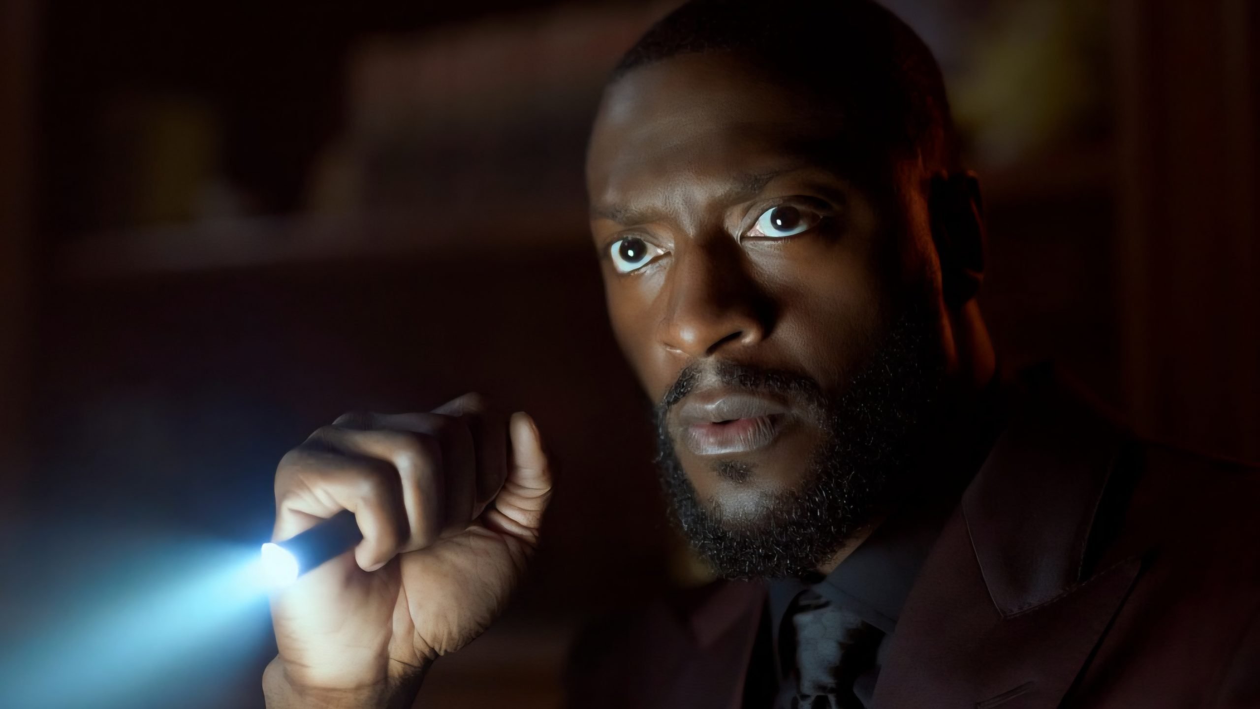 A man in a dark suit holds a flashlight, shining its beam ahead, with a serious expression in a dimly lit setting.