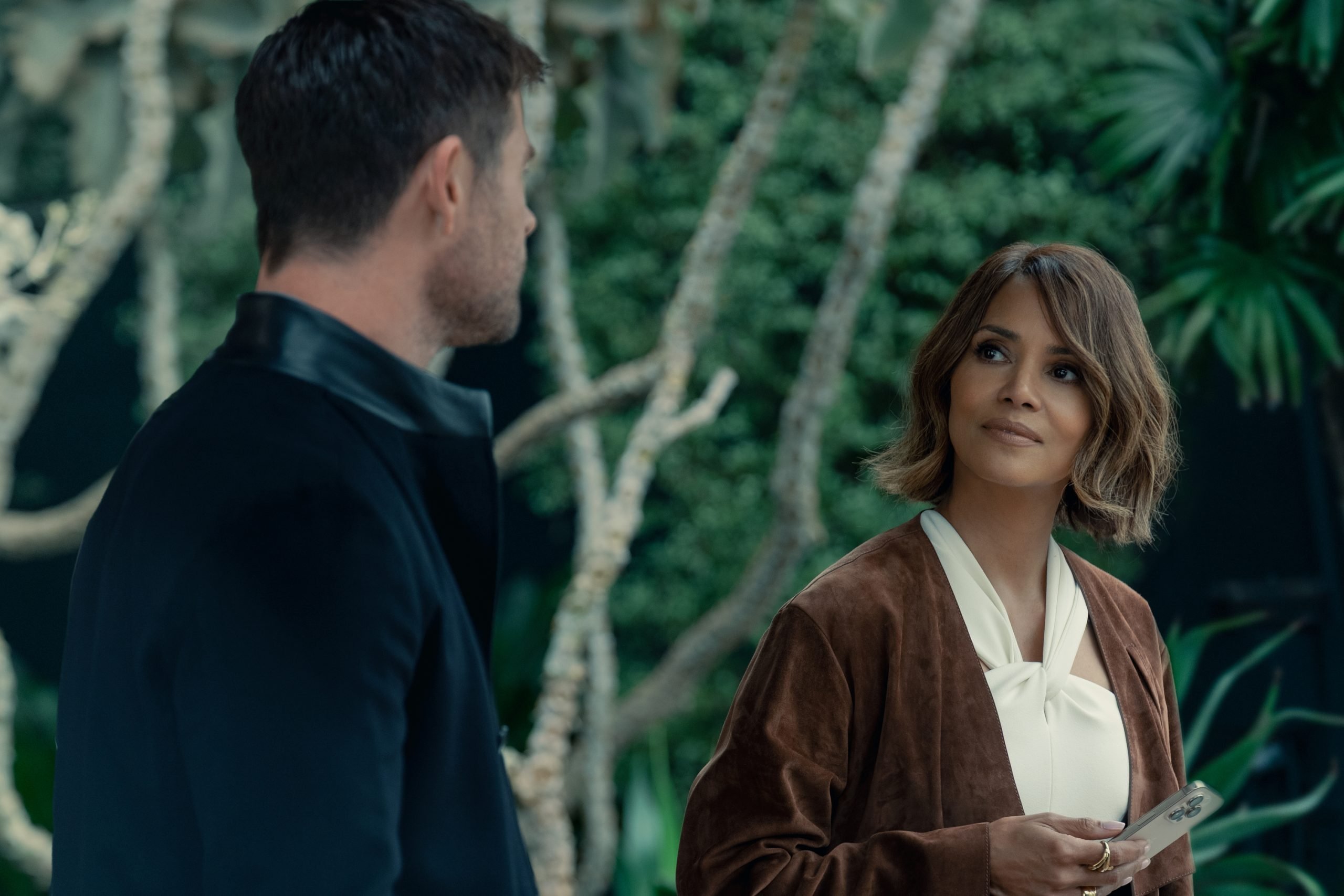 A woman holding a card looks at a man in a dark jacket; they are standing in front of greenery.