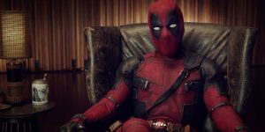 A person dressed in a red and black Deadpool costume sits in a brown leather armchair, with a lamp and a decorative jar on a side table nearby.