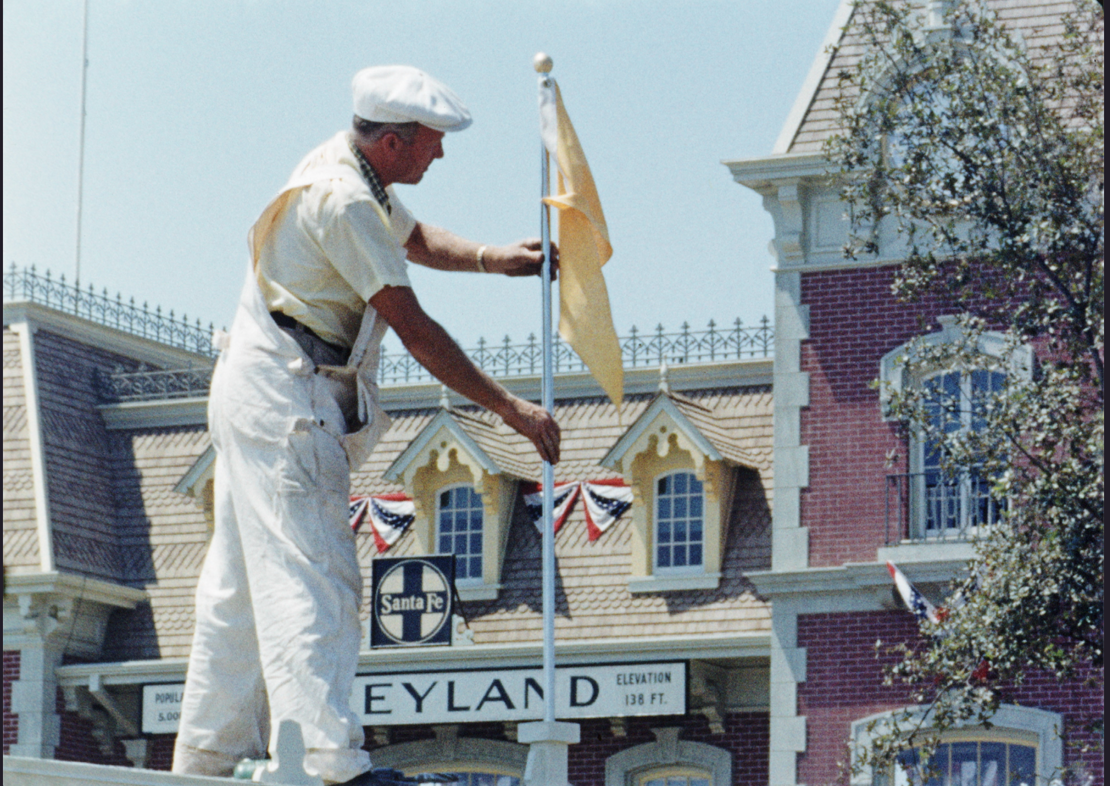A man in white work clothes raises a yellow flag on a pole in front of a Disneyland building decorated with red, white, and blue banners.