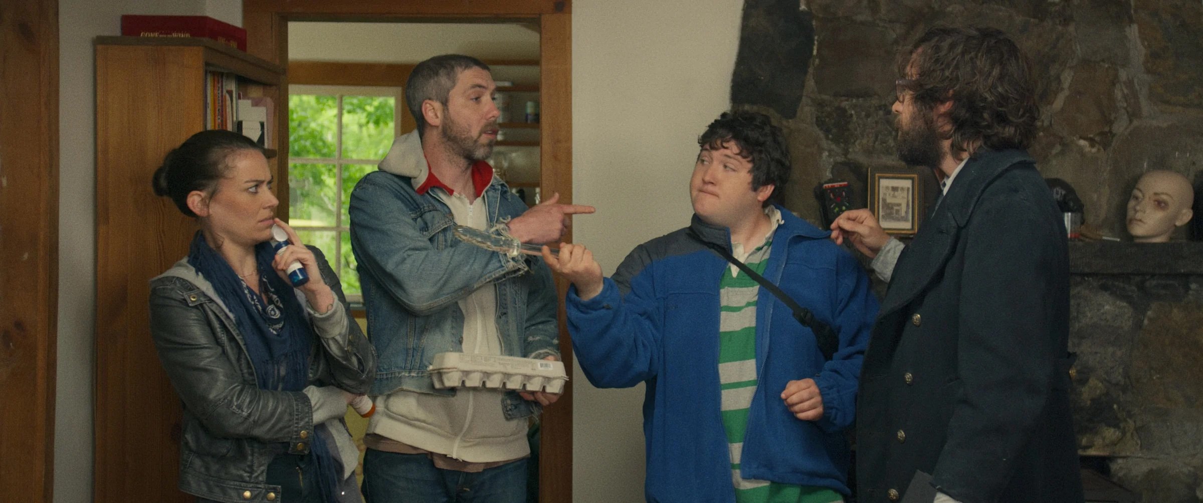 Four people are talking inside a room; one holds a carton of eggs, another points, and a mannequin head is visible on a shelf in the background.