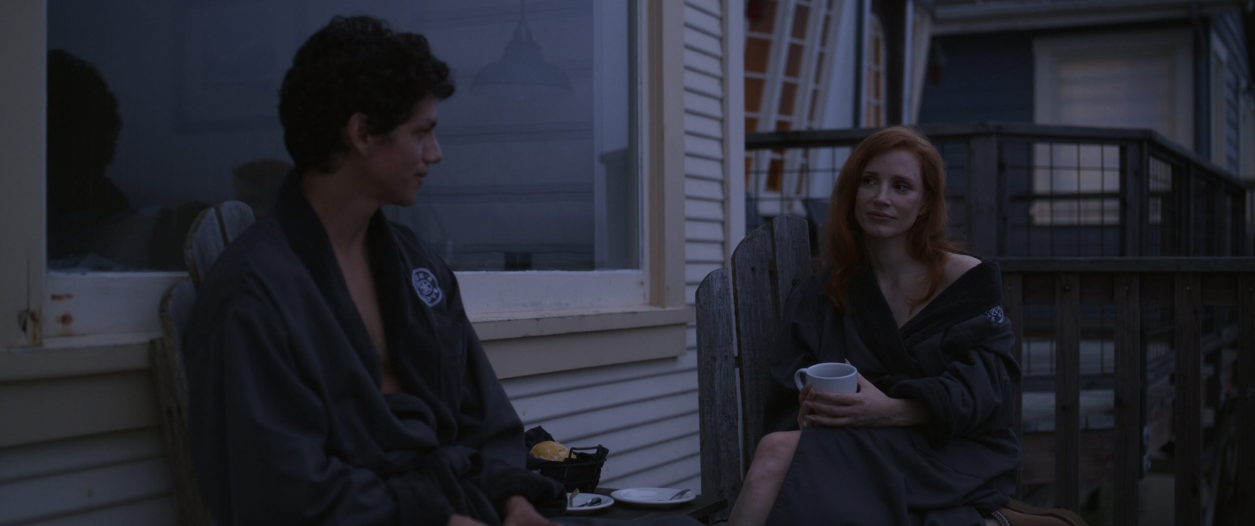 Two people in dark robes sit outside on wooden chairs near a house, drinking from mugs and talking in the early evening.