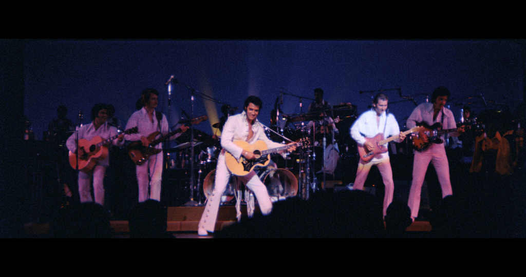A band dressed in white suits performs on stage with guitars and drums under blue lighting.