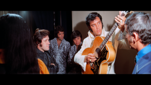 A young man playing an acoustic guitar during a live performance or rehearsal with a group of people.