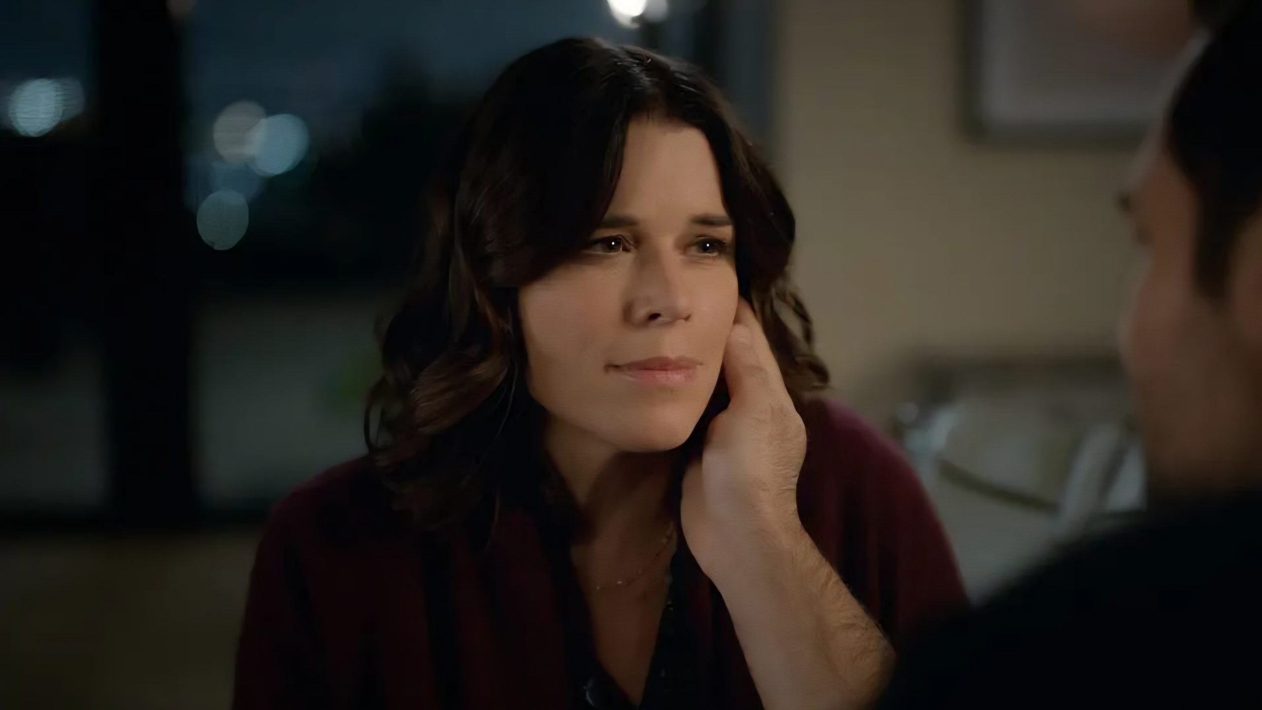 A person with shoulder-length dark hair sits indoors, looking thoughtfully at someone who is gently touching their face.