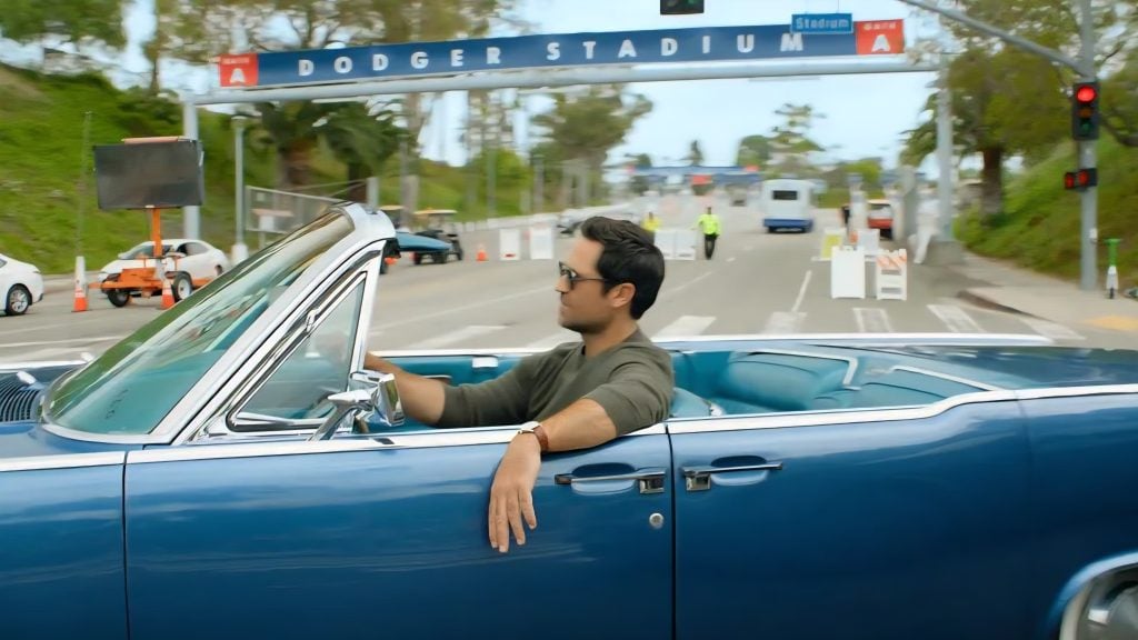 A man drives a blue convertible past the entrance sign for Dodger Stadium on a sunny day, with traffic cones and workers visible in the background.