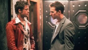 Two men stand facing each other indoors; one wears a red leather jacket and patterned shirt, the other a gray suit jacket and white shirt.