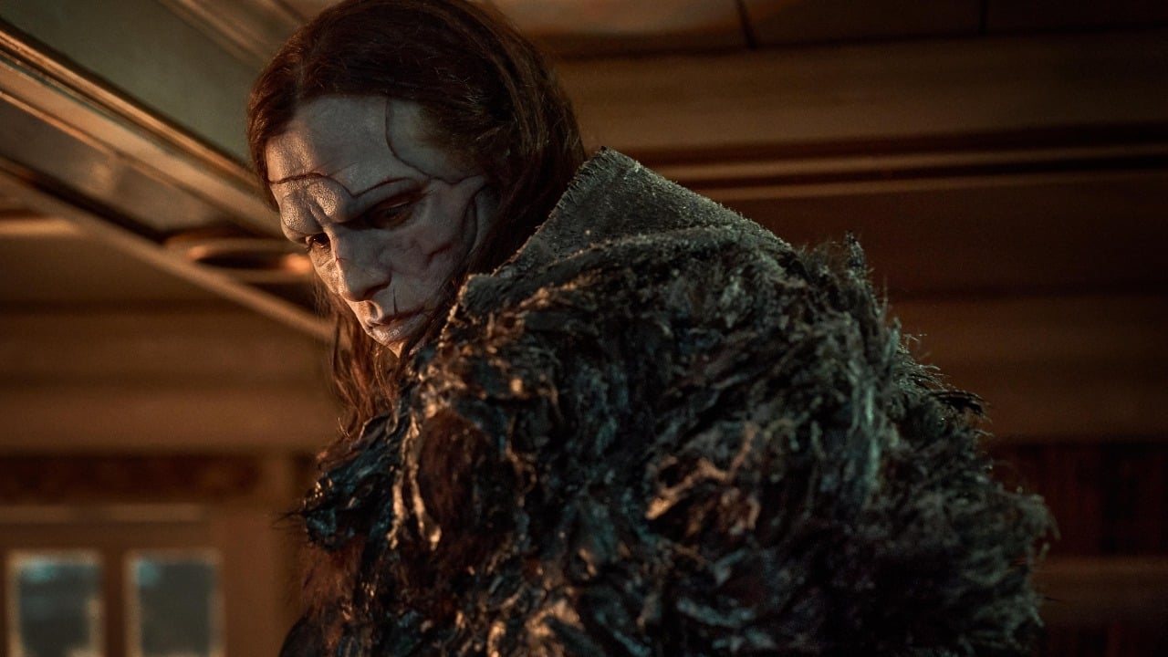 A person with long dark hair and a pale, heavily scarred face wears a tattered, dark, textured cloak in a dimly lit room.