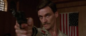 A man in a military uniform points a handgun directly forward inside a room with an American flag in the background.