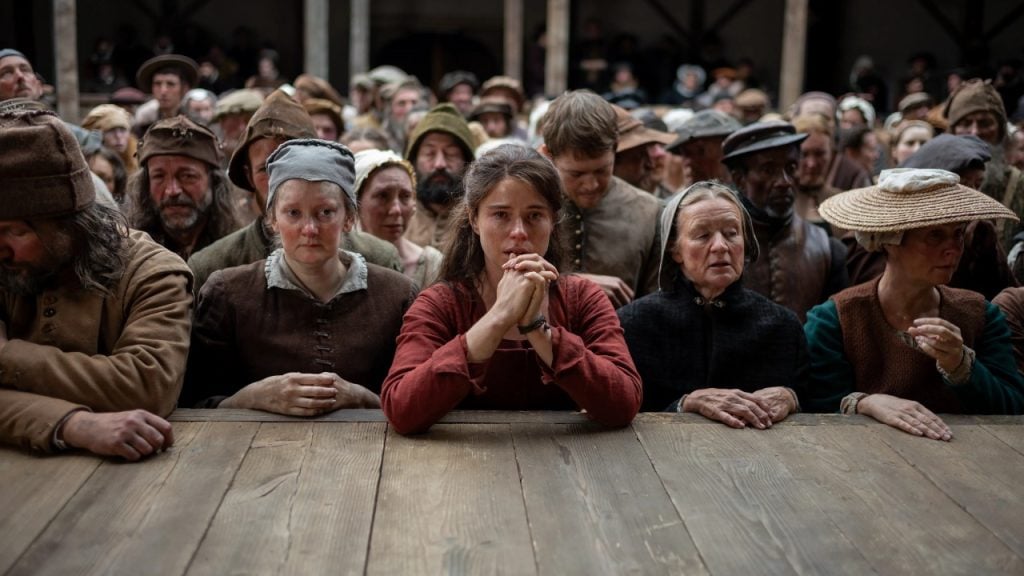 A group of people in historical clothing stand behind a wooden barrier, looking somber and focused, with a young woman at the center clasping her hands.