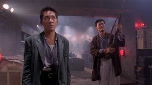 Two men stand in a dim, industrial room; one looks ahead with a serious expression while the other holds a shotgun and looks to the side.