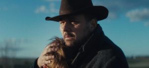 A man in a dark coat and wide-brimmed hat embraces a person with light brown hair outdoors under a blue sky.