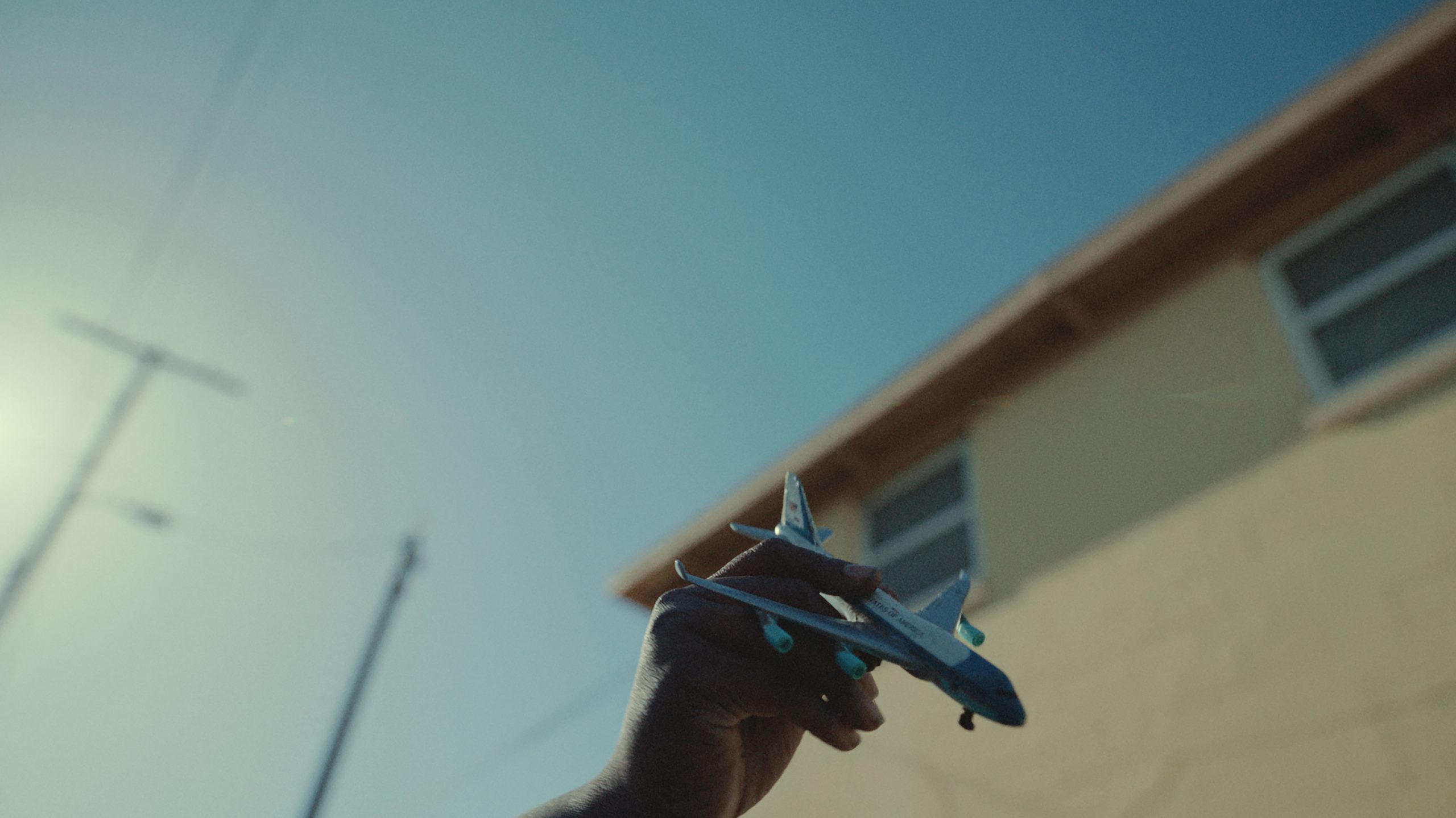A hand holds a small toy airplane up against the blue sky, with part of a building and a utility pole visible in the background.