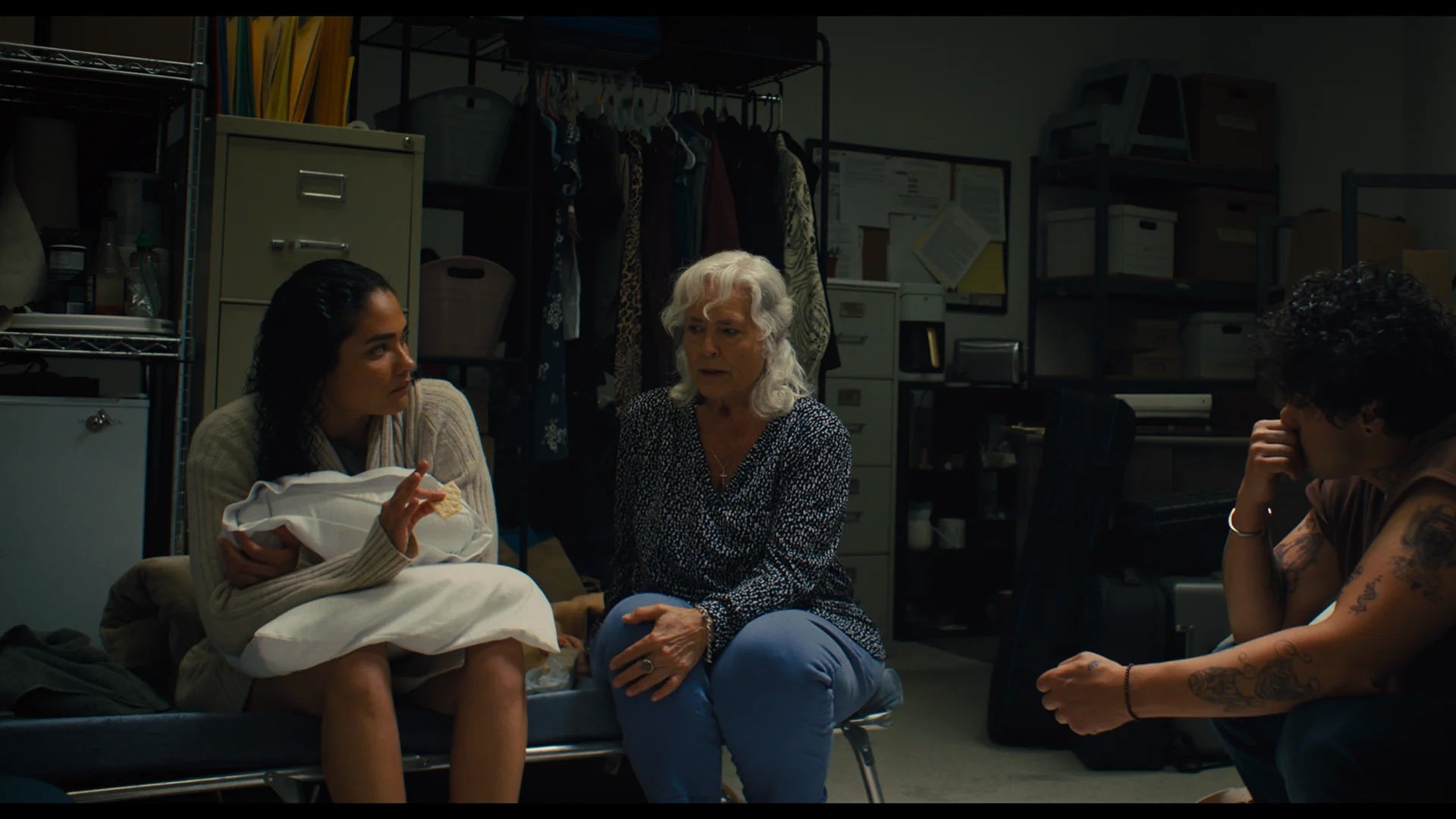 Three people sit in a storage room, engaged in a serious conversation; shelves, boxes, and a clothing rack are visible in the background.