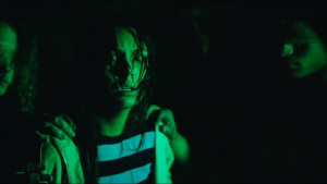 A person with a distressed expression is illuminated by green light, with hands on their shoulders and shadowy figures in the dark background.