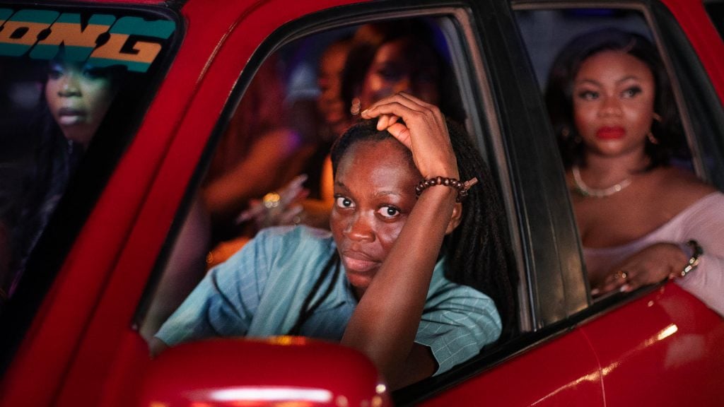 A person in a blue shirt sits in the driver's seat of a red car, resting their head on their hand, with several passengers in the back seat at night.