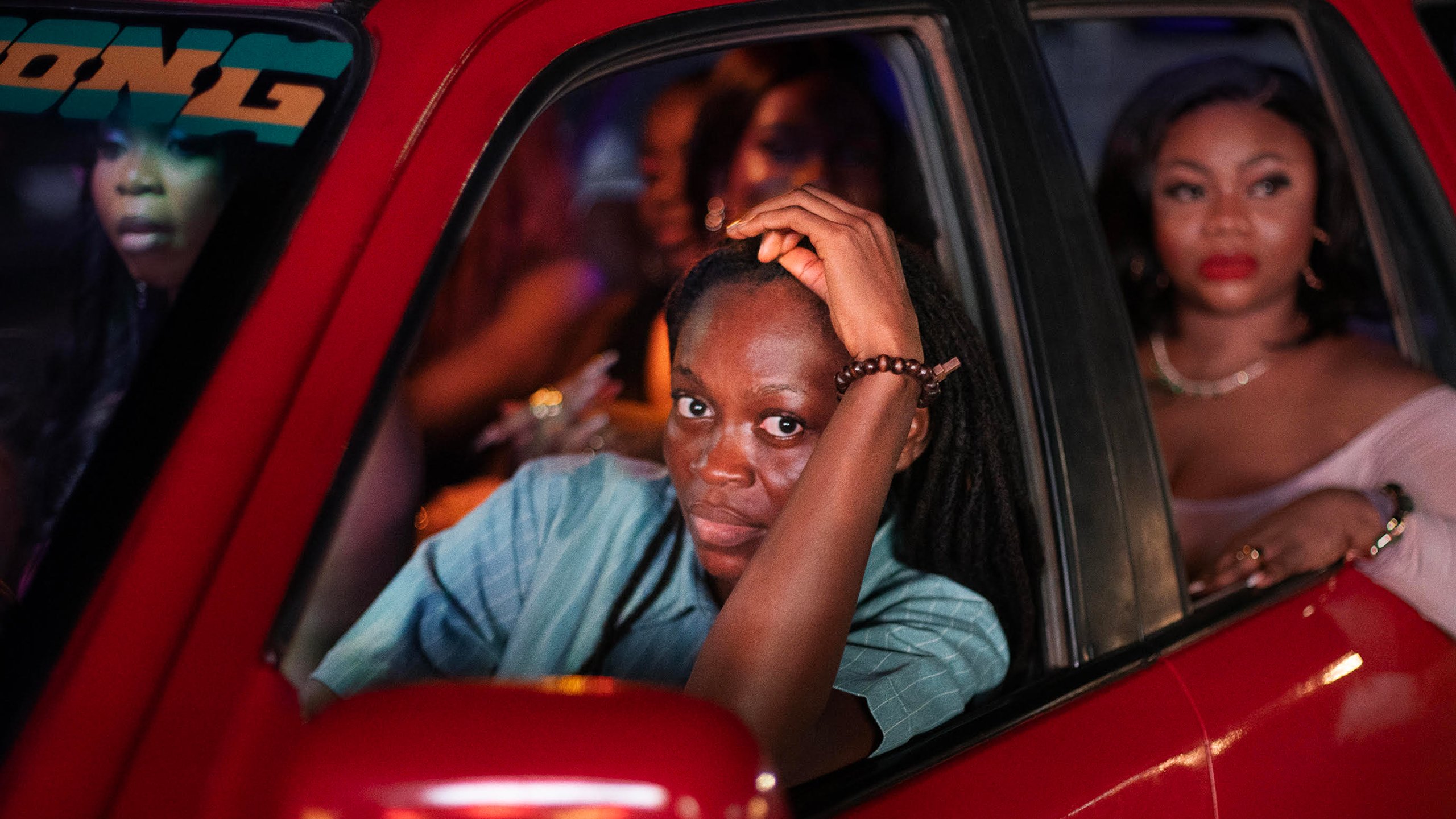 A person in a blue shirt sits in the driver's seat of a red car, resting their head on their hand, with several passengers in the back seat at night.