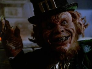A person wearing leprechaun makeup and costume, with pointed ears, sharp teeth, and a black hat, appears to smile menacingly in a dimly lit setting.