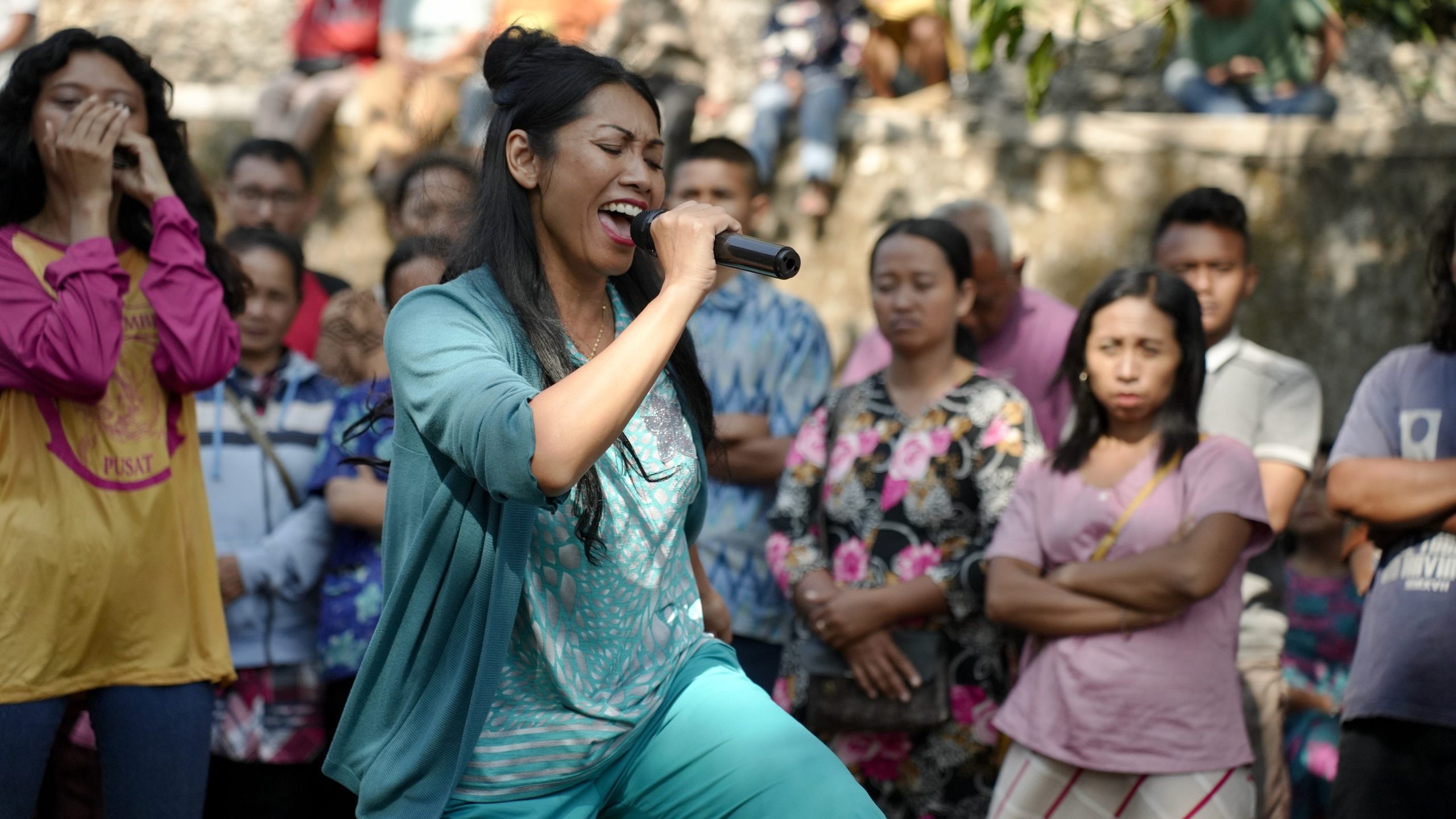 A woman sings passionately into a microphone in front of a crowd of people outdoors, with some audience members listening and others watching.