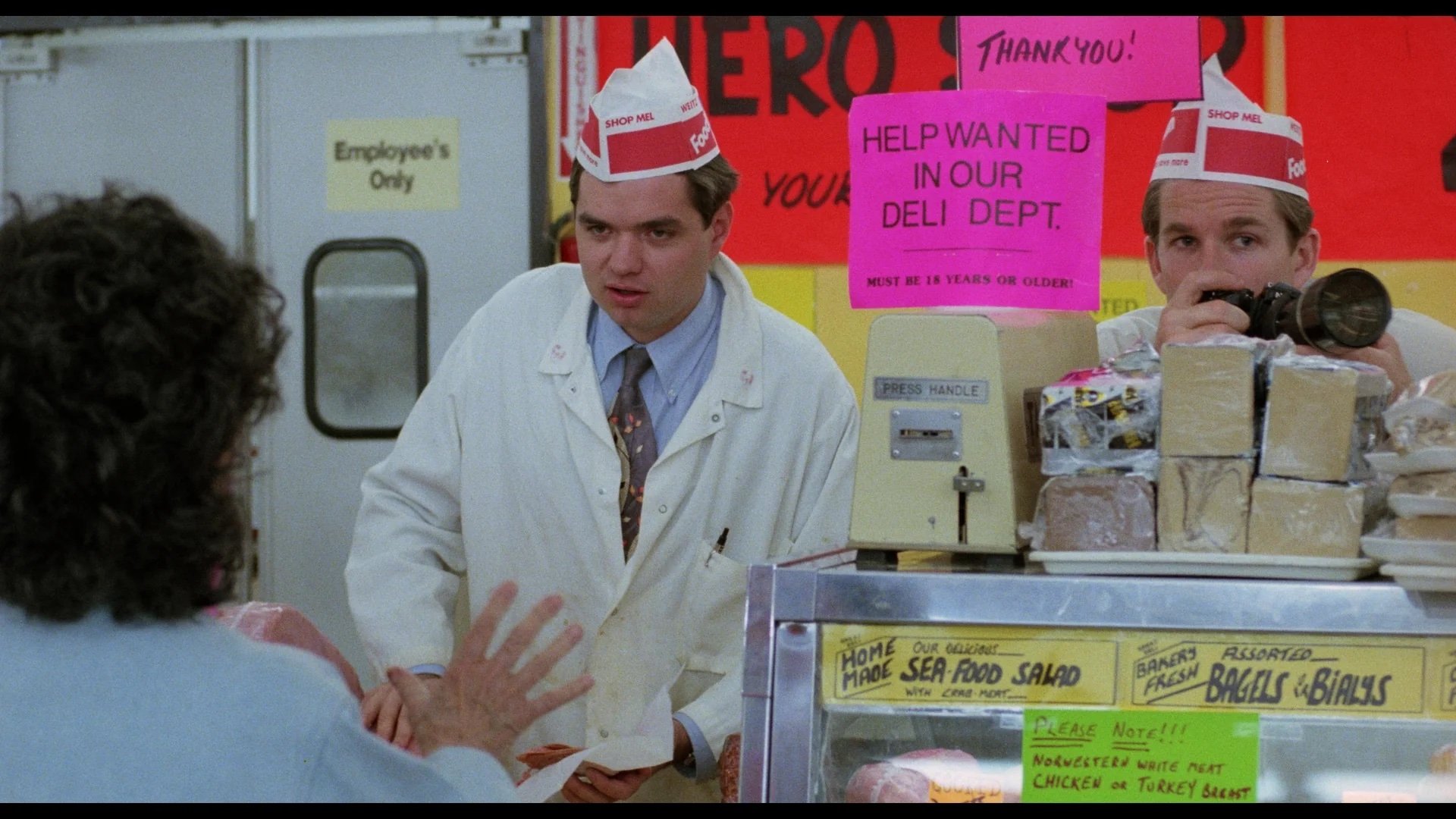 Two deli workers in white coats and red hats stand behind a counter with deli meats and signs, talking to a customer. One worker is writing, the other holds a microphone.