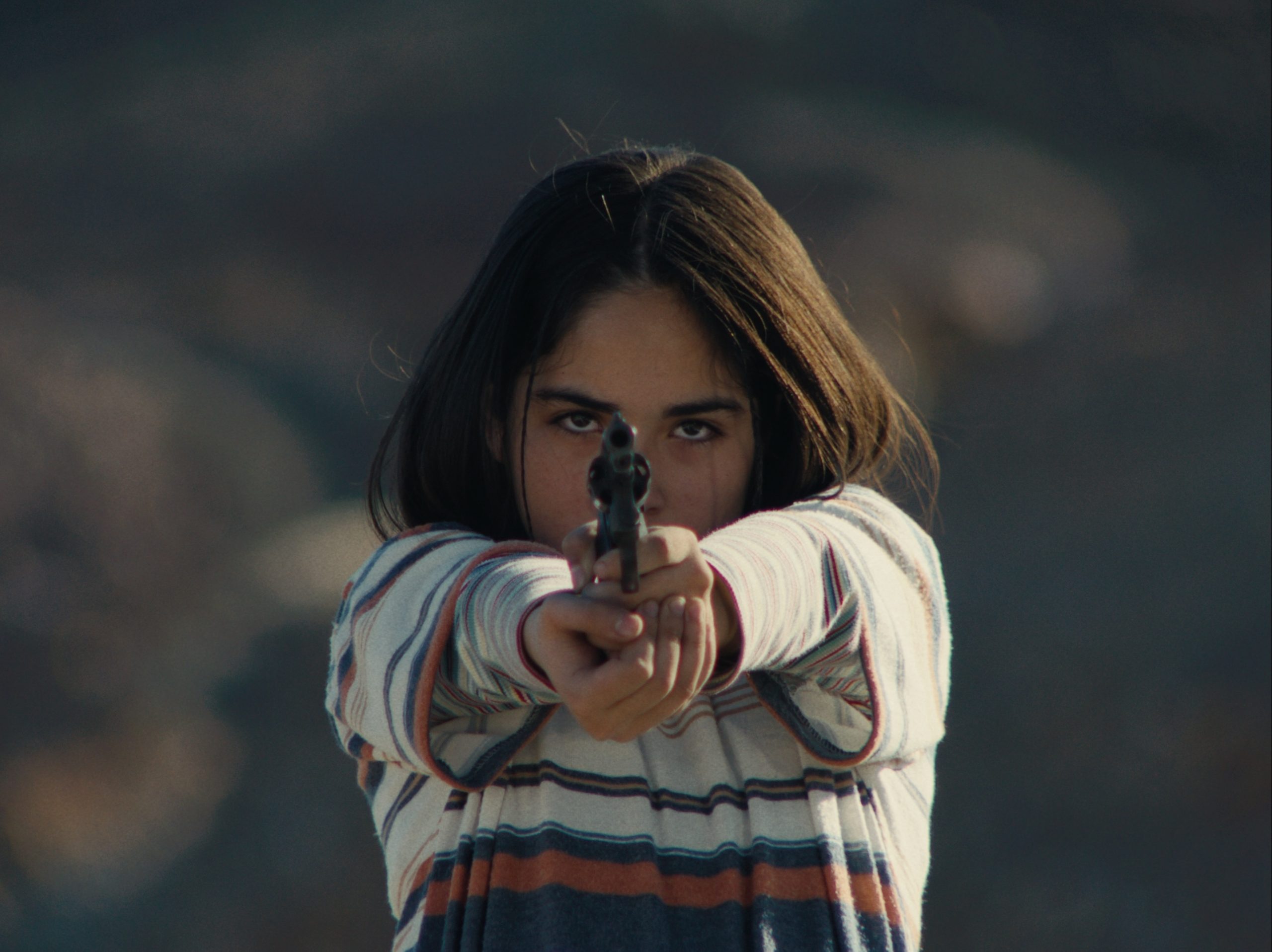 A young girl with long dark hair and a striped shirt points a handgun directly at the camera, standing outdoors with a blurred background.
