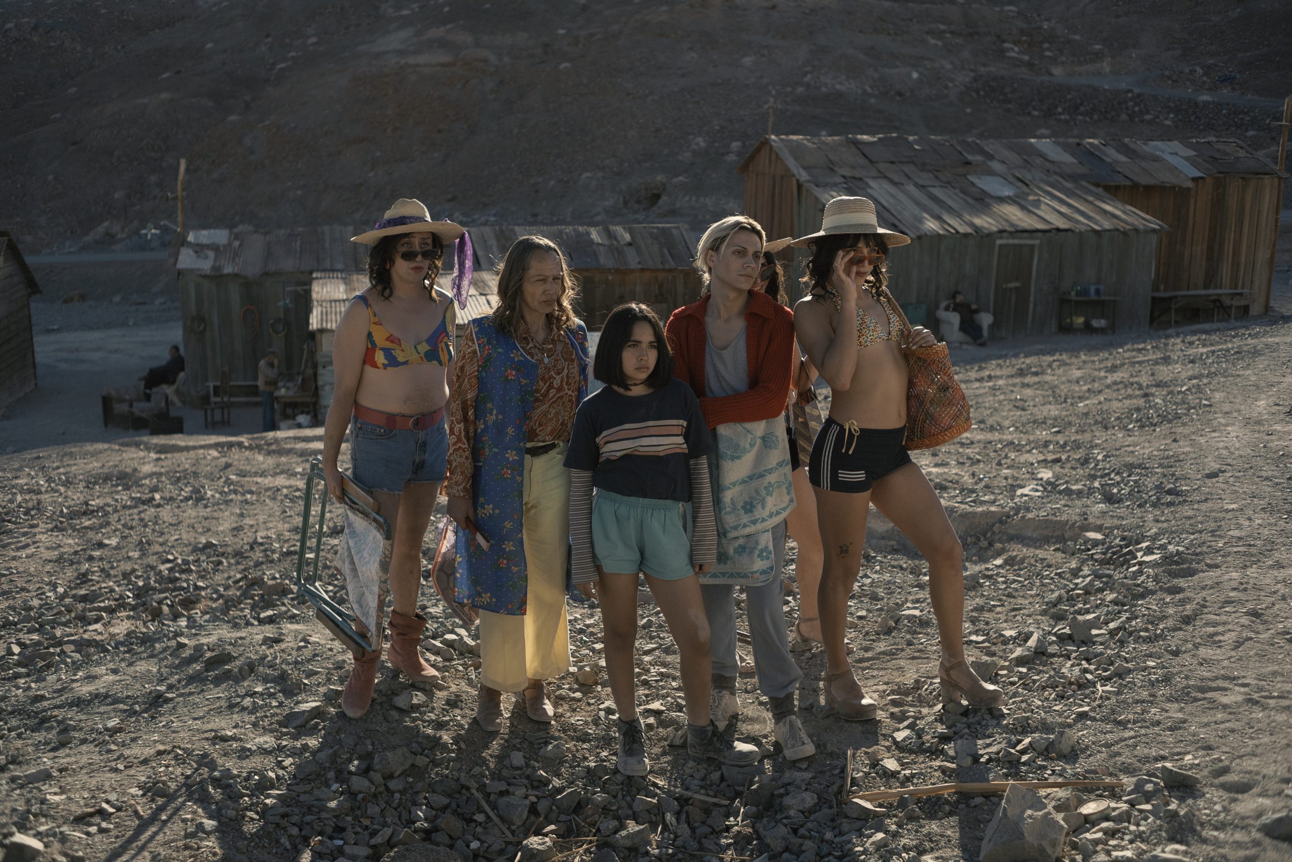 Five people stand together on rocky ground in front of rustic wooden buildings, wearing casual and colorful summer clothes in a rural, barren landscape.