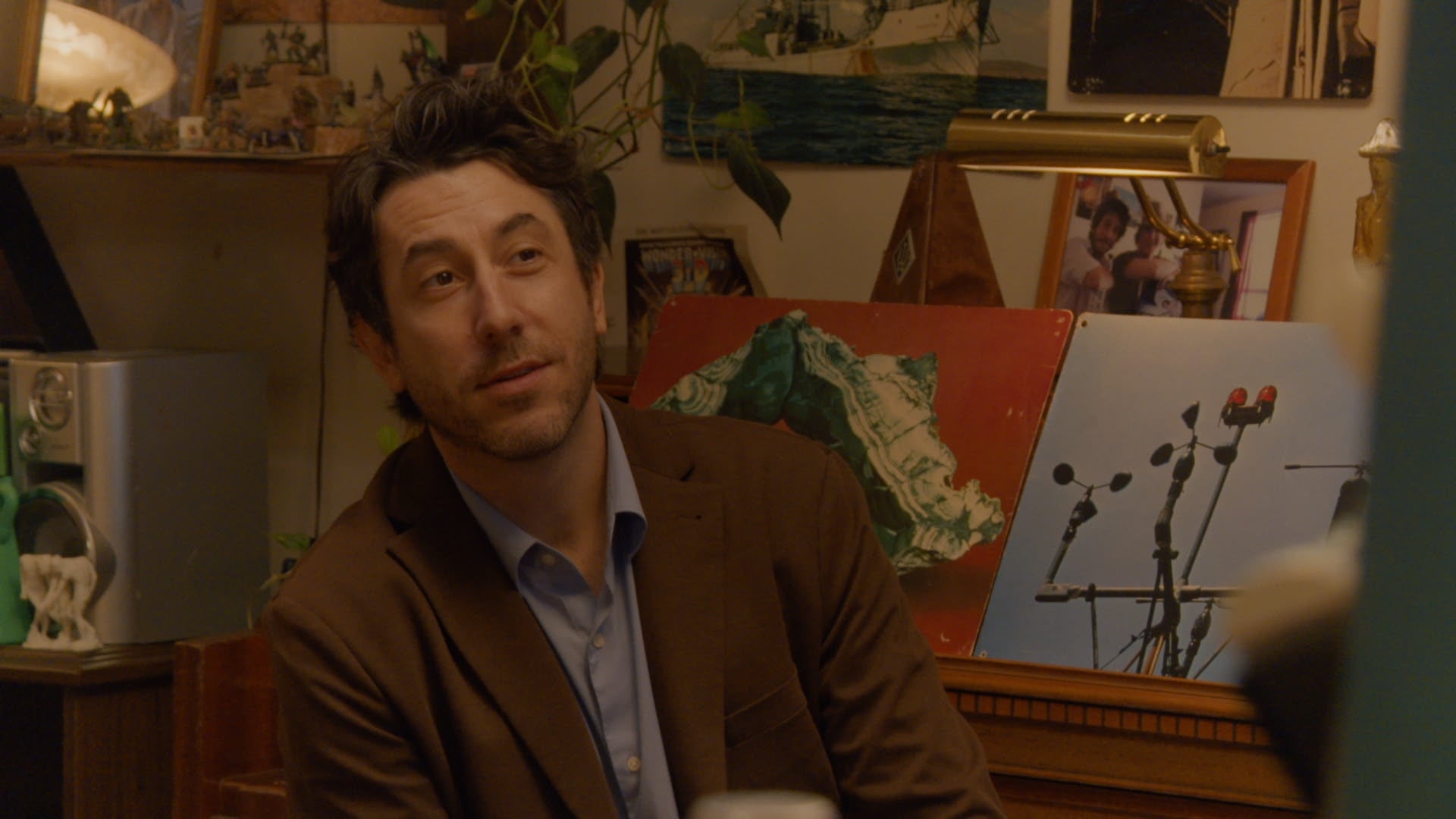 A man in a brown blazer sits indoors, with framed photos, plants, and artwork on a piano behind him.