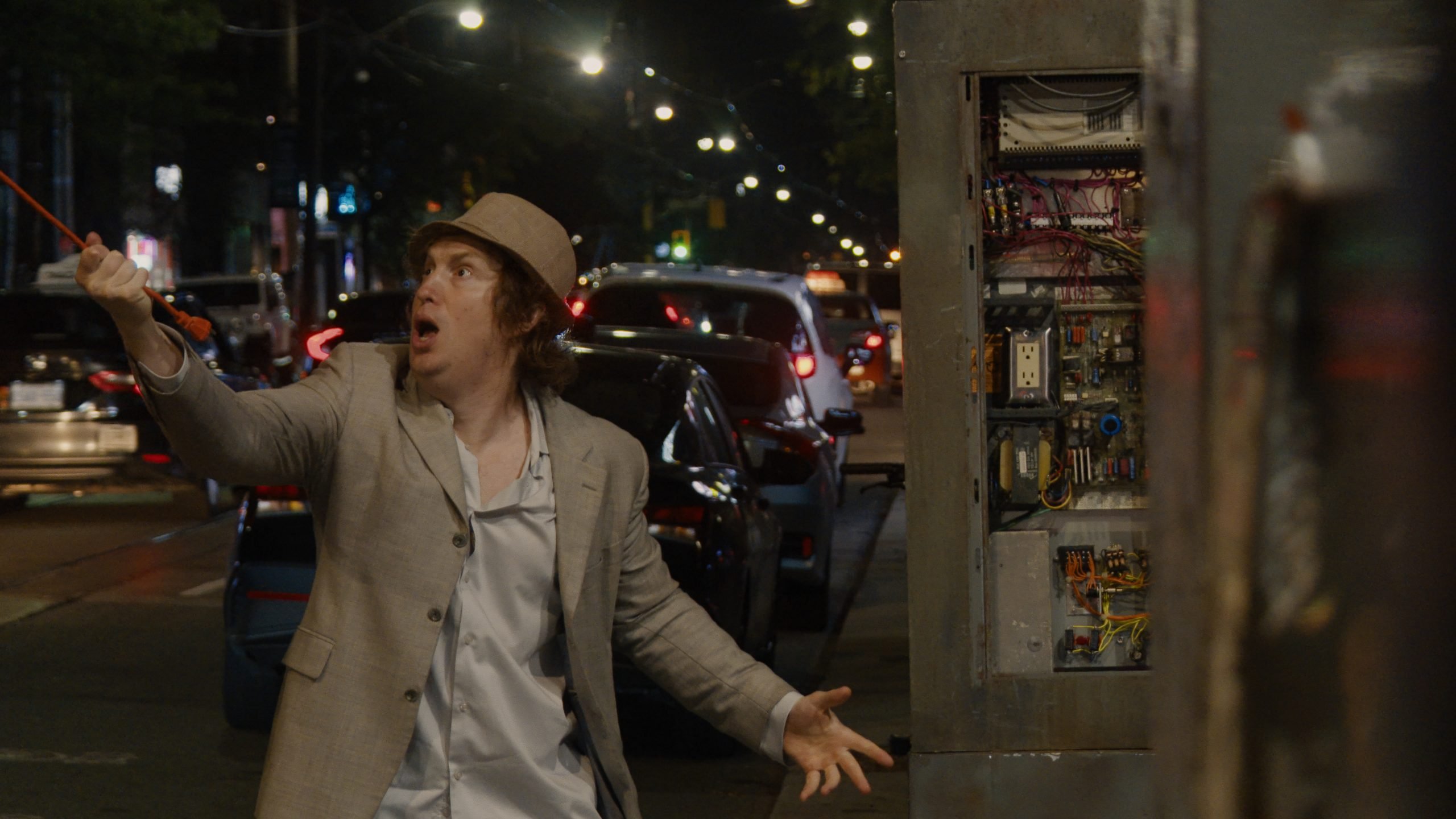 A man in a light suit and hat stands on a city street at night, holding a red leash, facing an open electrical panel with a surprised expression.