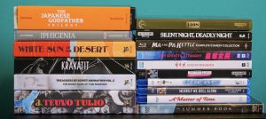 A stack of DVD and Blu-ray movie cases including titles like "The Japanese Godfather Trilogy," "Iphigenia," "Silent Night, Deadly Night," and "The Summer Book," against a teal background.