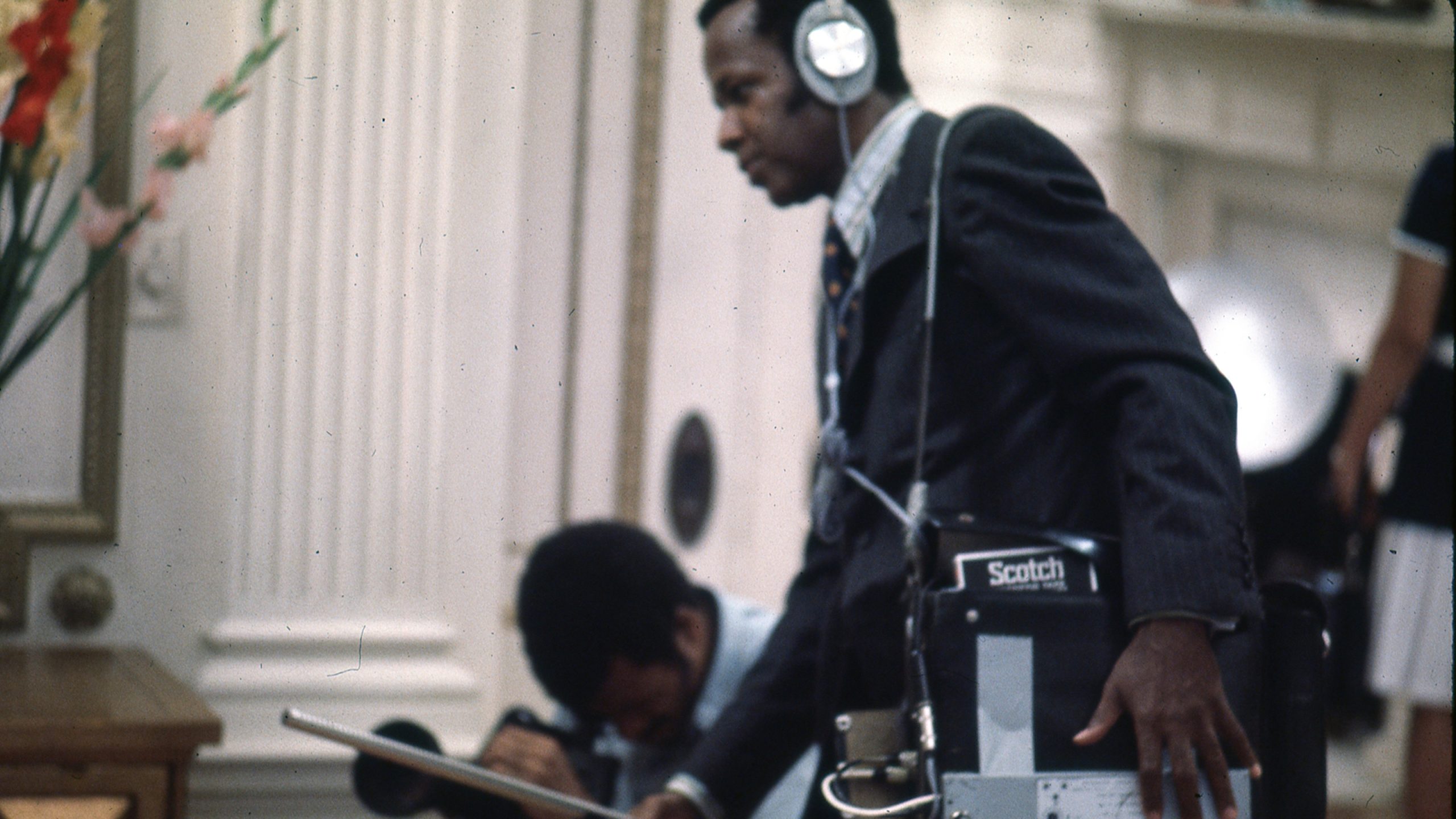 A man in a suit operates audio equipment with headphones on, holding a reel-to-reel tape recorder labeled "Scotch." Another person crouches nearby, adjusting equipment.