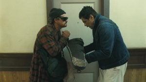 Two men stand in a hallway in front of a closed door; one holds a large ski boot while the other stands with hands clasped, wearing dark glasses and a beanie.