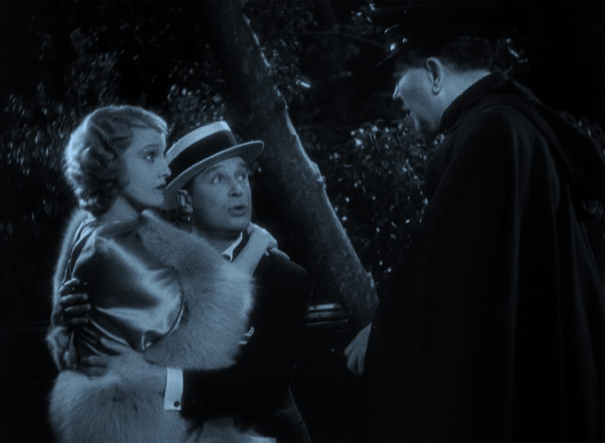 A man holds a woman in his arms while speaking to another man in a cloak and hat at night, with trees in the background.
