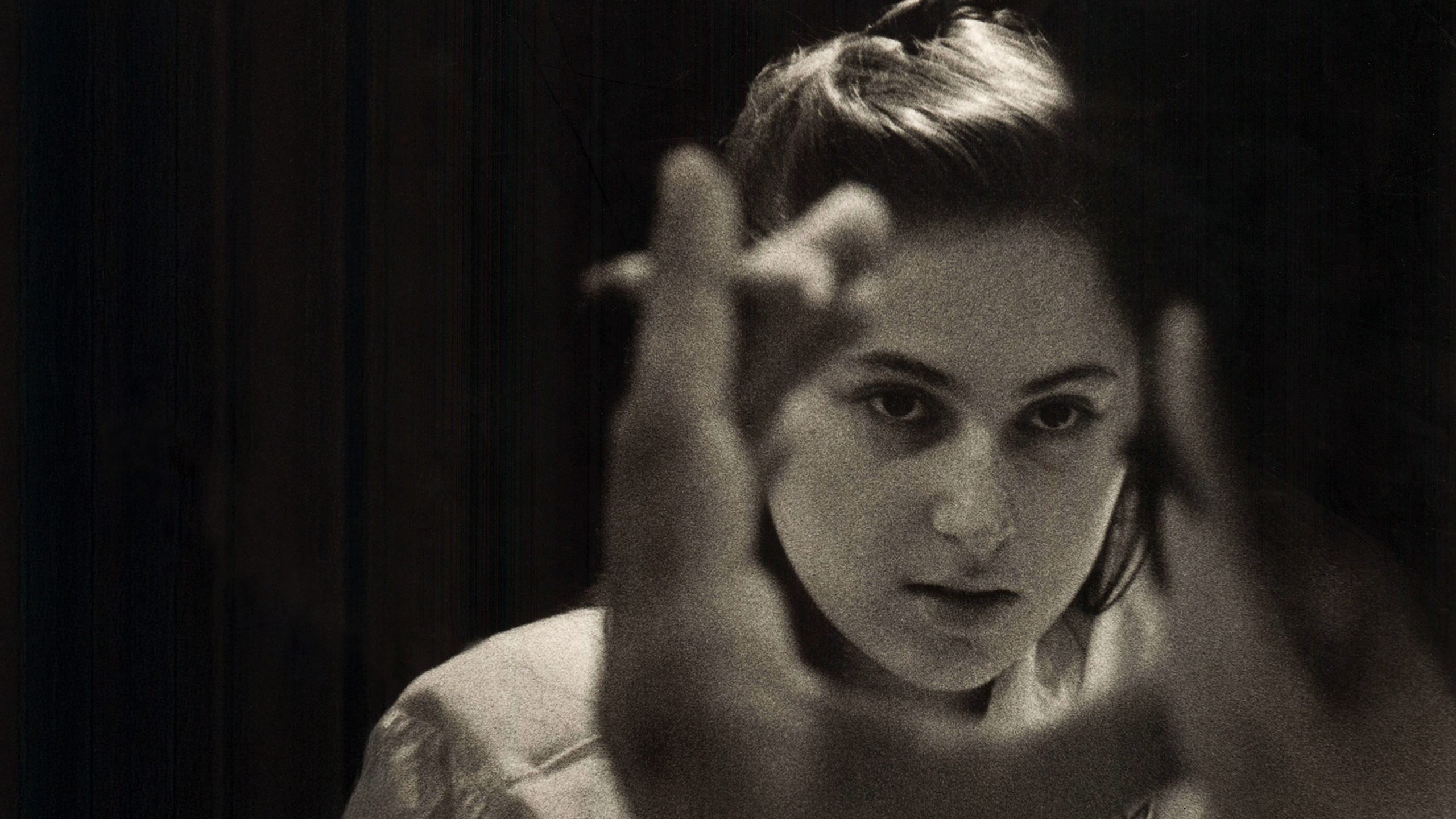 A young woman with a serious expression looks through her hands, which are positioned in a circular frame in front of her face. The image is in black and white.