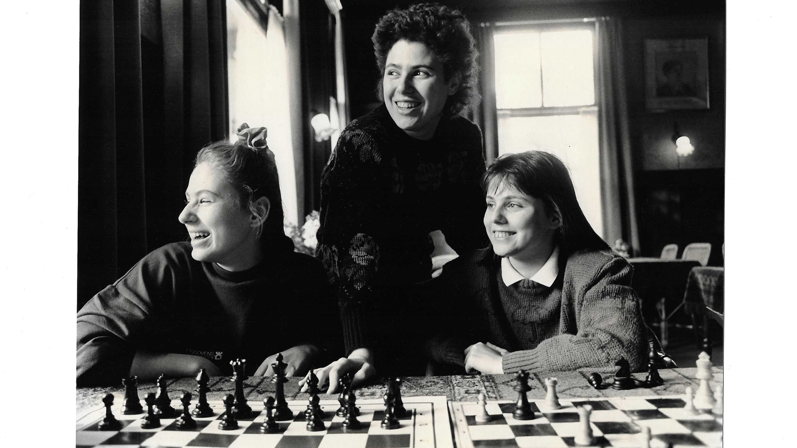 Three women sit at a table with chess boards, smiling and looking in different directions in a well-lit room.