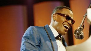 A man in a blue suit and sunglasses sings passionately into a vintage microphone while playing a white piano.