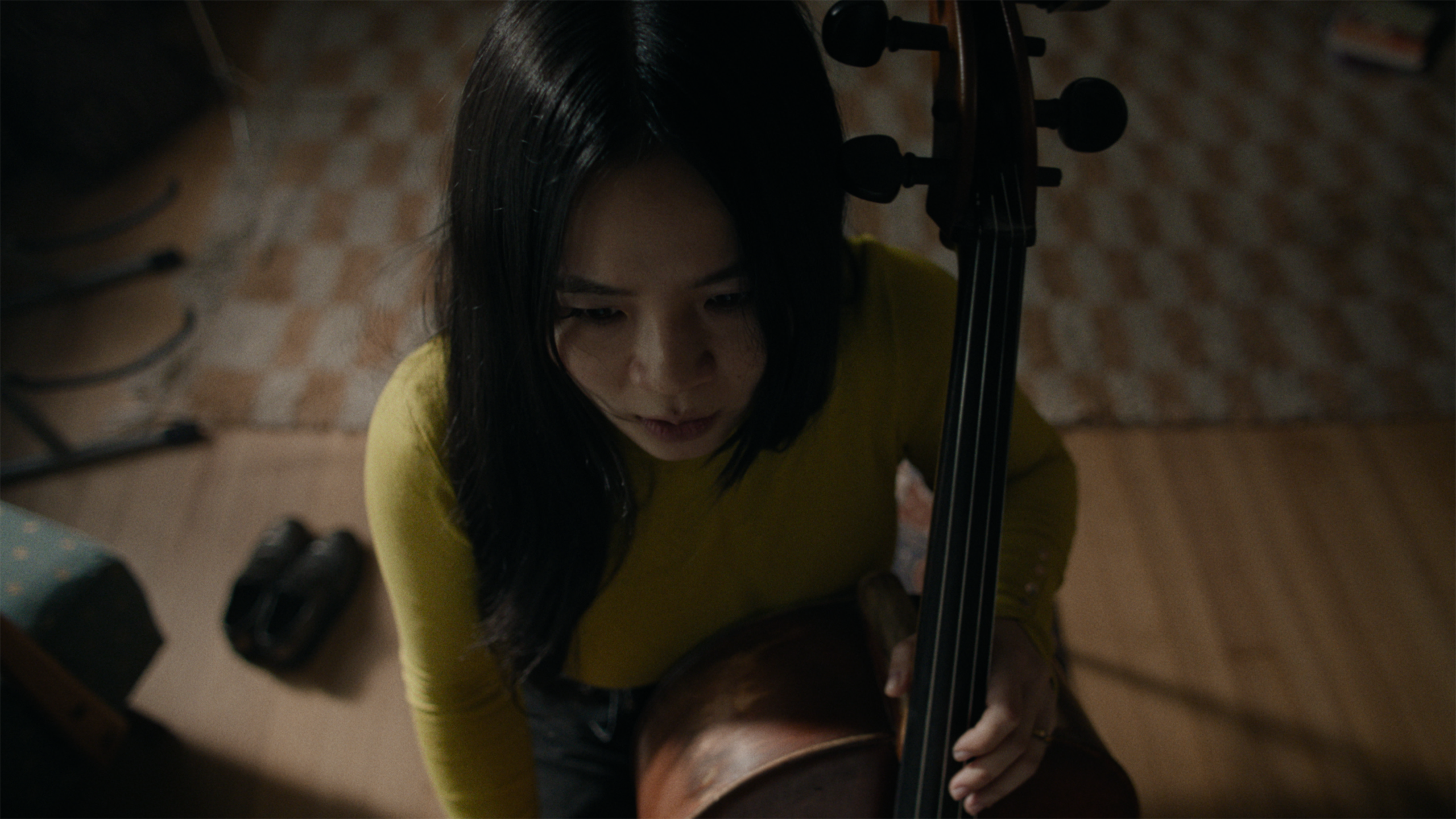 A woman with long dark hair in a yellow top holds a cello, viewed from above in a dimly lit room with a patterned rug and wooden floor.
