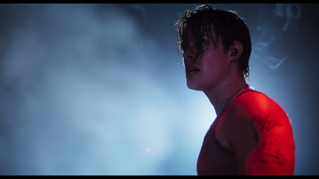 A person wearing a sleeveless red shirt and earpiece stands in profile on a smoky stage, lit from behind with blue light.