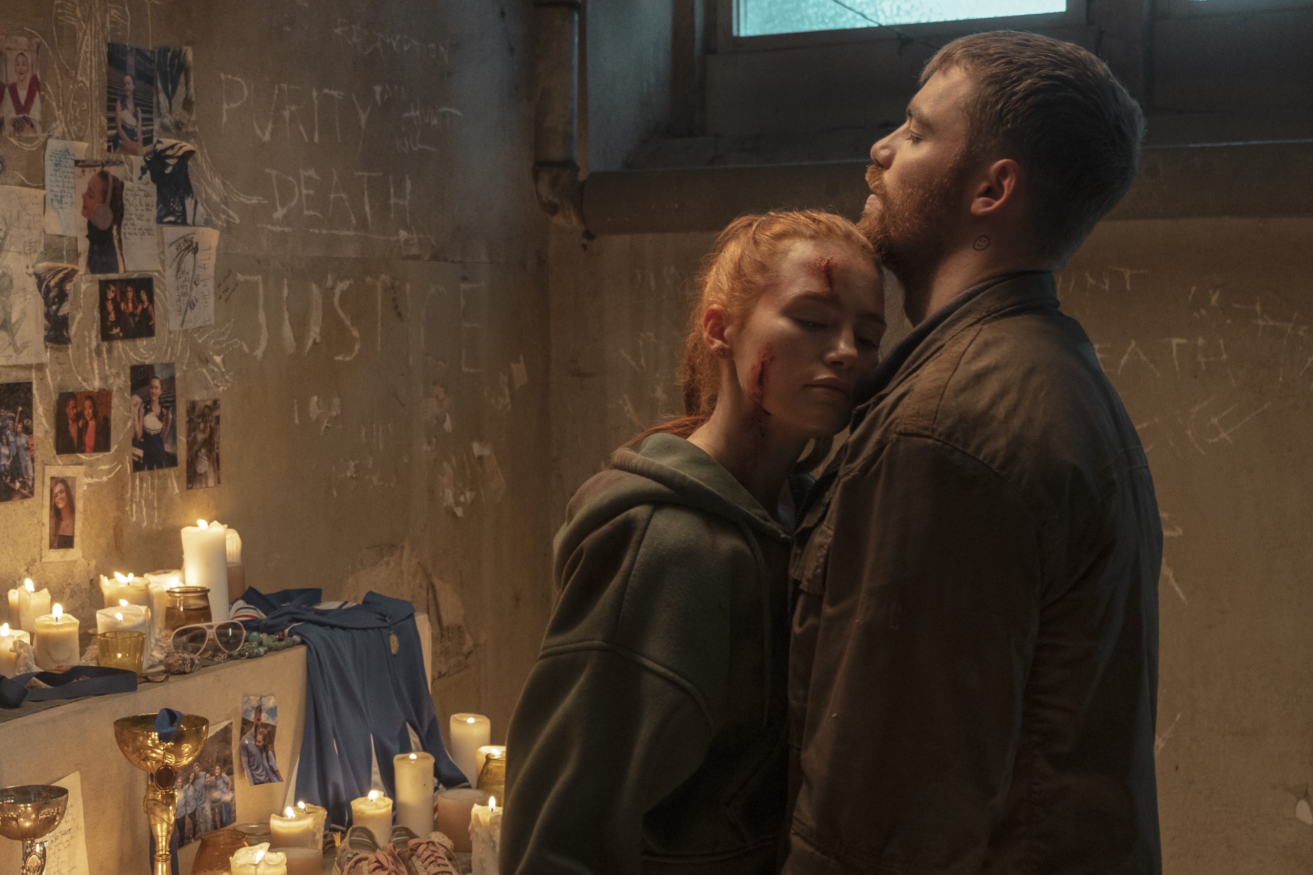 A woman with a bloody forehead rests her head on a man's chest in a dimly lit room with candles and photos on the wall behind them.