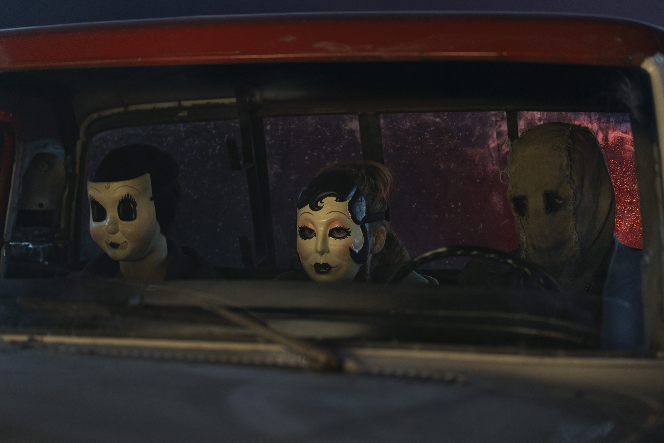 Three people wearing eerie masks sit in the front seat of an old vehicle at night, seen through the windshield.